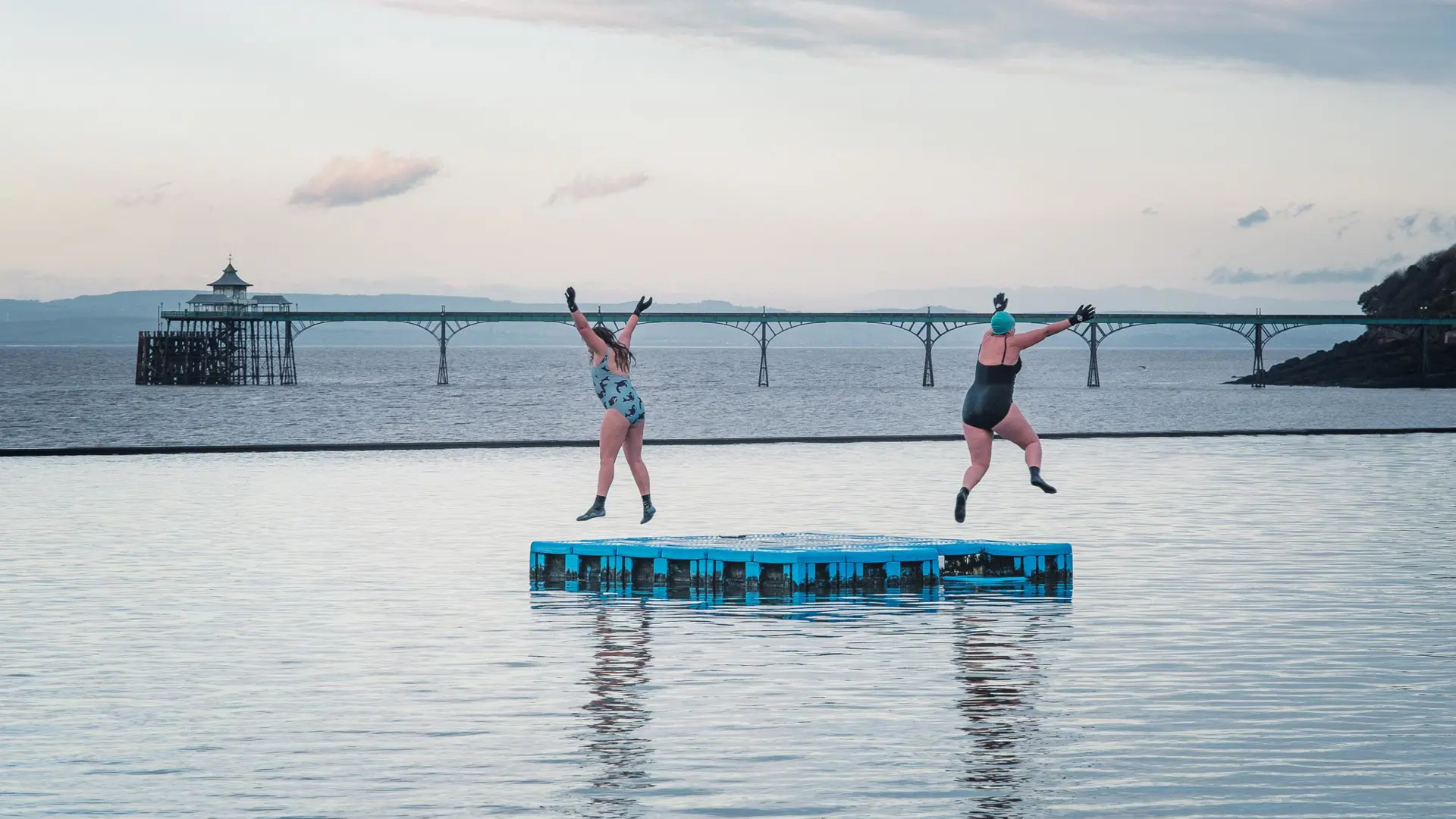 Two ladies jumping off the marine lake pontoon in Clevedon, overlooking Clevedon pier