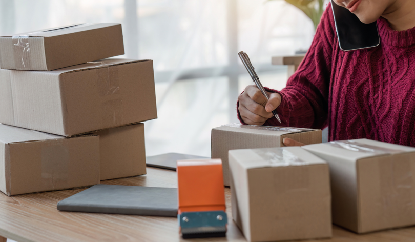 A lady writing an address on a parcel whilst talking on the phone surrounded by multiple packages